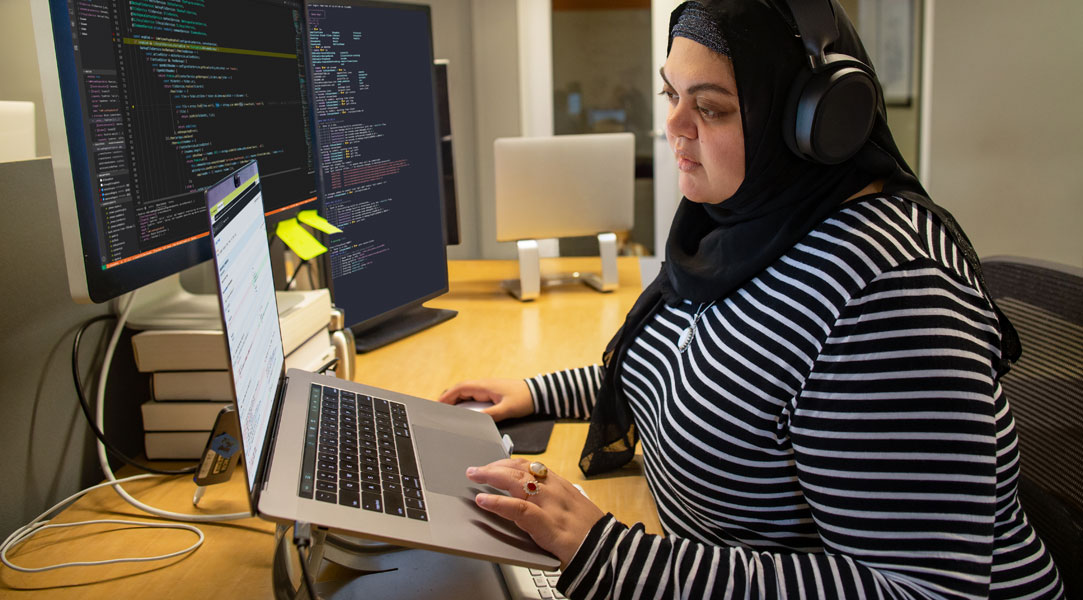 Eine Entwicklerin, die an ihrem Schreibtisch programmiert, Hijab und Kopfhörer trägt, hat ihren Arbeitsplatz mit einer Einrichtung mit mehreren Monitoren angepasst.